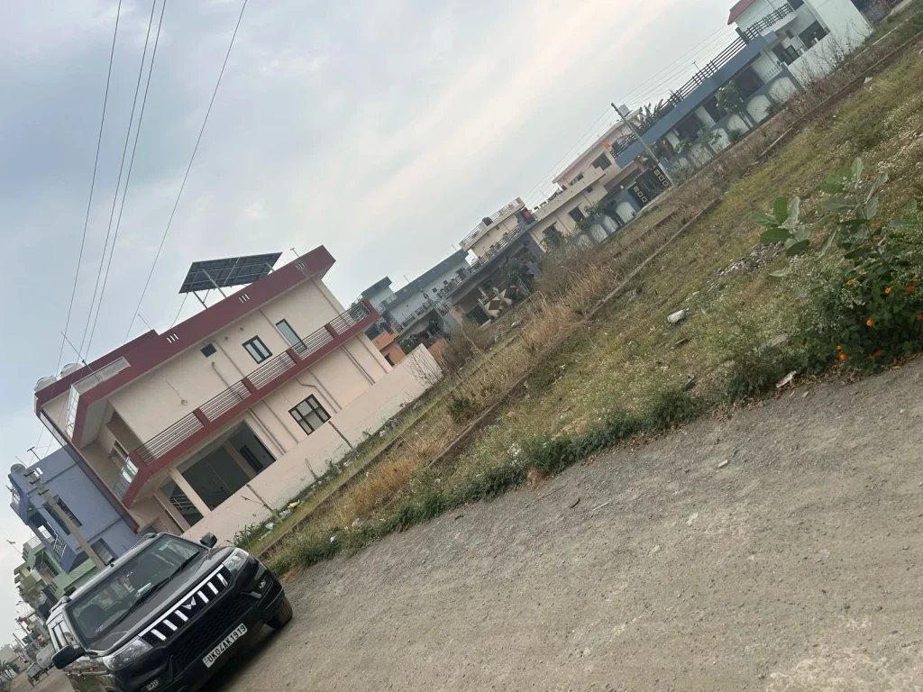 Empty plot beside a white boundary wall, with trees in the distance and an overcast sky in a residential zone of Haldwani
