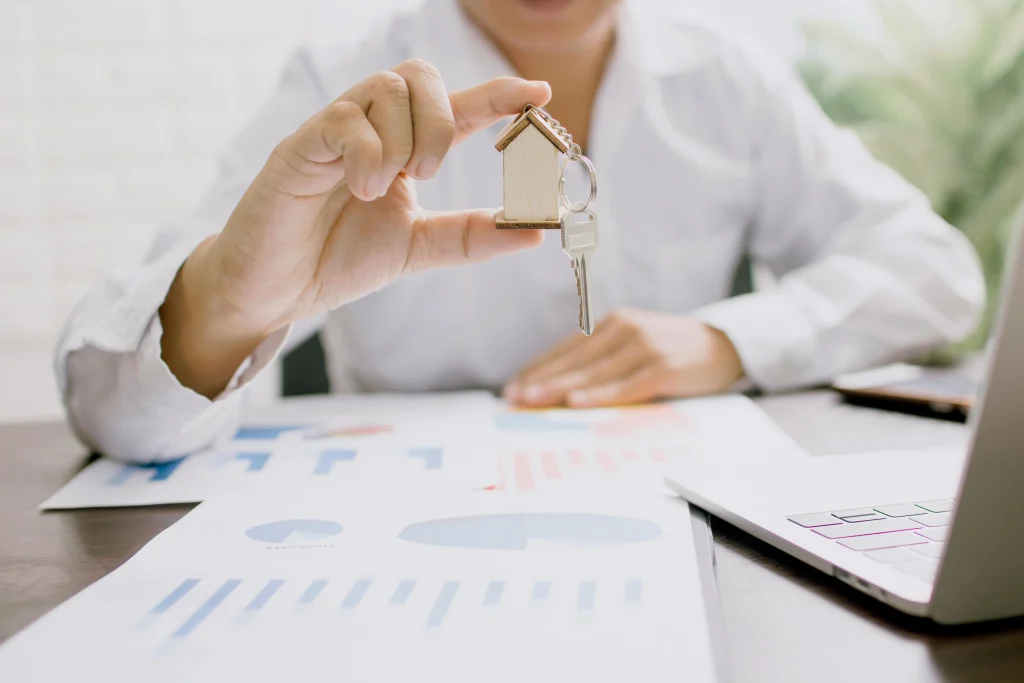 Real estate agent holding house keychain with key, financial charts and laptop on desk