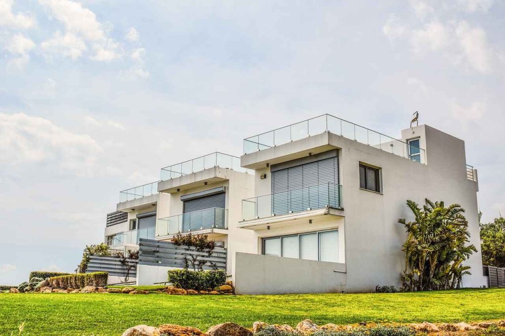 Modern white duplex house with large glass balconies and a well-maintained green lawn under a partly cloudy sky.